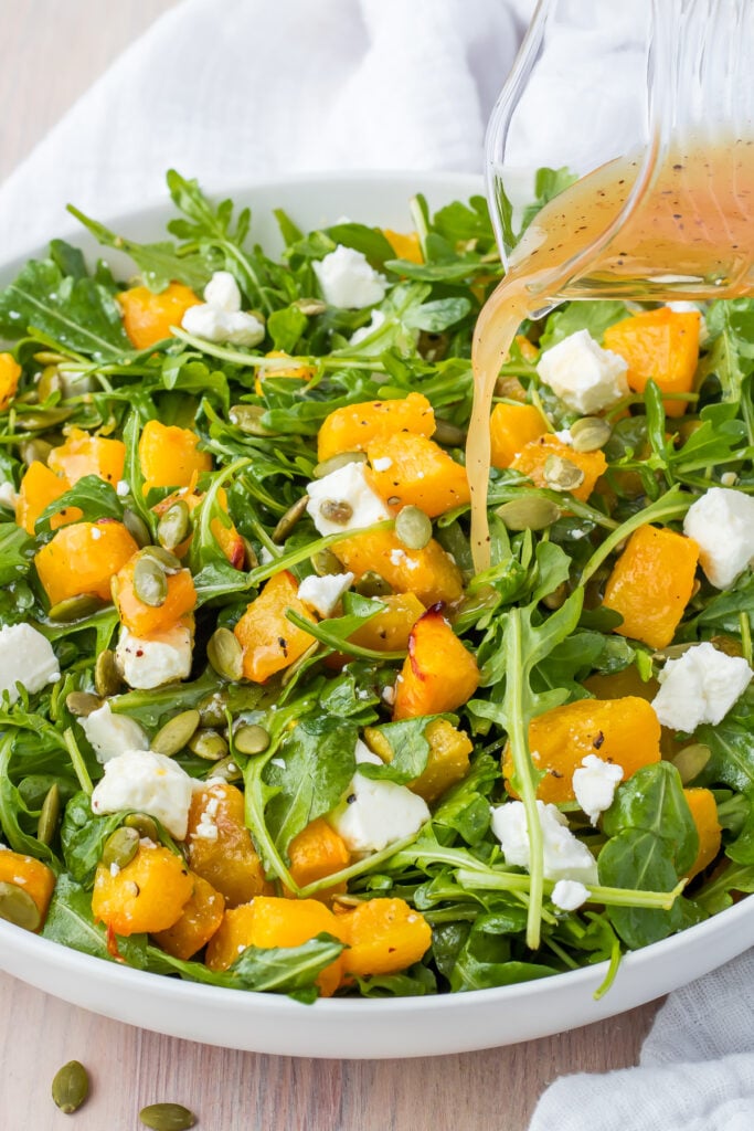 A fresh salad with arugula, roasted butternut squash, crumbled feta cheese, and pumpkin seeds, drizzled with vinaigrette from a glass pitcher. The vibrant dish is served in a white bowl on a wooden table with a white cloth.