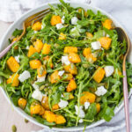 A fresh roast pumpkin salad with arugula, roasted pumpkin, crumbled feta cheese, and pumpkin seeds, served in a white bowl. A gold spoon and fork are placed on the side, with a white napkin underneath.