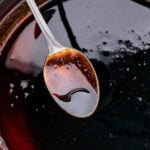 Close-up of a silver spoon dipped in dark, glossy balsamic glaze. The liquid boasts a rich, deep color and smooth texture with small bubbles on the surface, beautifully reflecting light.