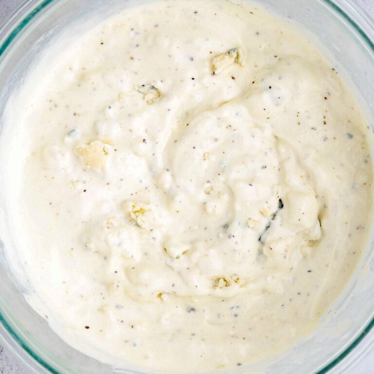 A glass bowl filled with creamy blue cheese dressing. The dressing has visible chunks of blue cheese and is speckled with black pepper. The texture appears smooth and thick.