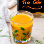 A glass brimming with bright orange fire cider, adorned with painted leaves, rests on a wooden table. Behind it stands a jar labeled "Fire Cider." Nearby, a white cloth and sprigs of fresh rosemary complete the scene.