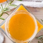 A top view of a glass jar filled with vibrant orange fire cider, set on a light wooden surface. Fresh rosemary sprigs and an orange are visible around the jar, with a white cloth in the background.
