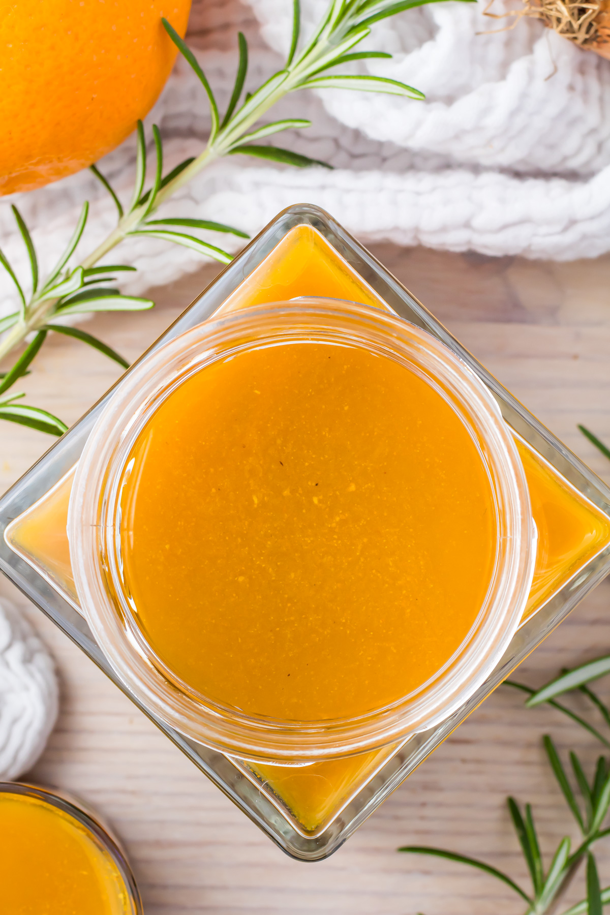 Top view of a square, glass jar filled with golden honey, surrounded by sprigs of fresh rosemary and part of an orange. A white cloth covers part of the light wooden surface.
