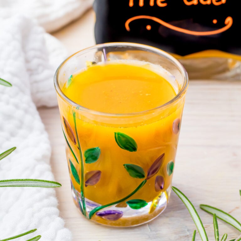 A glass filled with a vibrant orange juice, decorated with hand-painted green leaves and purple buds. The glass sits on a light wooden surface surrounded by sprigs of rosemary. A partially visible sign in the background.