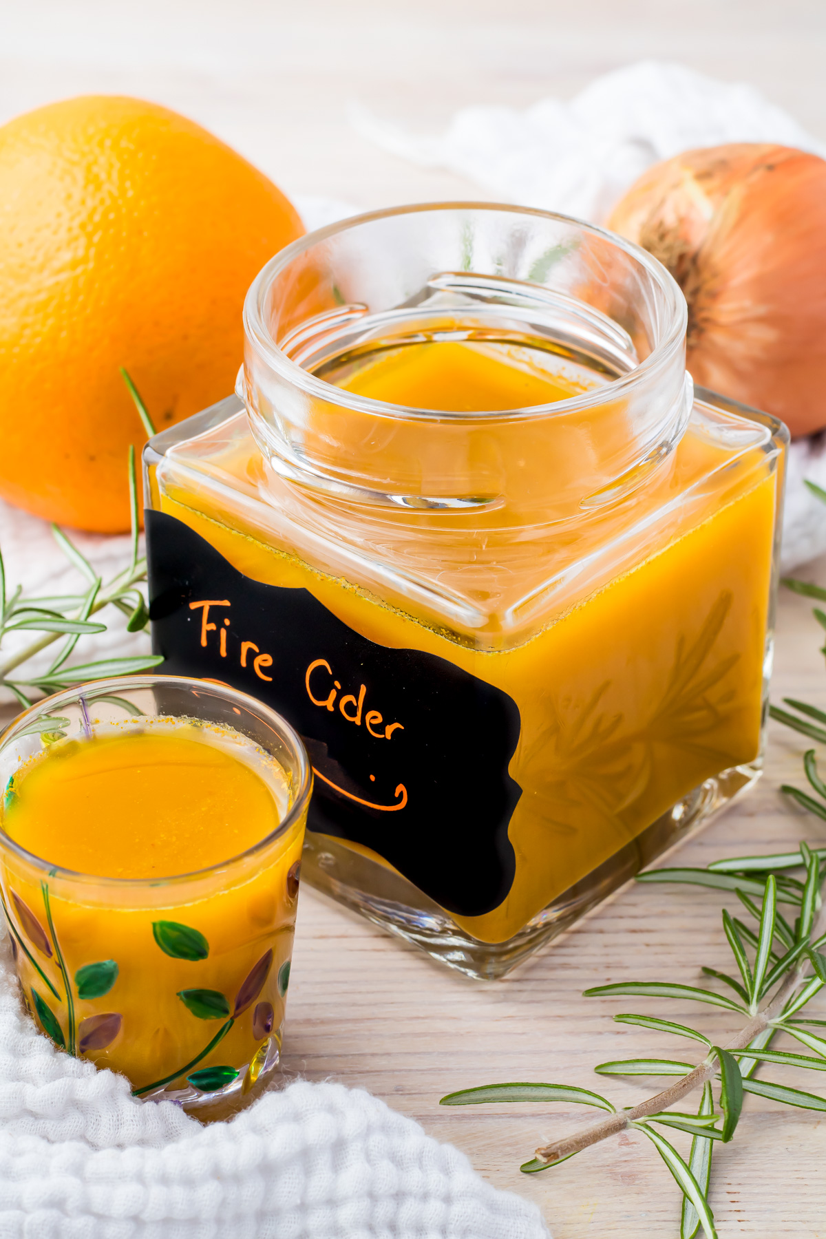 A glass jar labeled "Fire Cider" filled with an orange liquid. Next to it, a small glass with the same liquid. Surrounding the jar are an orange, an onion, and sprigs of rosemary on a light-colored surface.