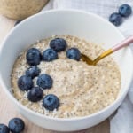 A bowl of keto overnight oats topped with fresh blueberries and shredded coconut, with a golden spoon resting inside. The bowl is placed on a light wooden surface with a white cloth nearby.