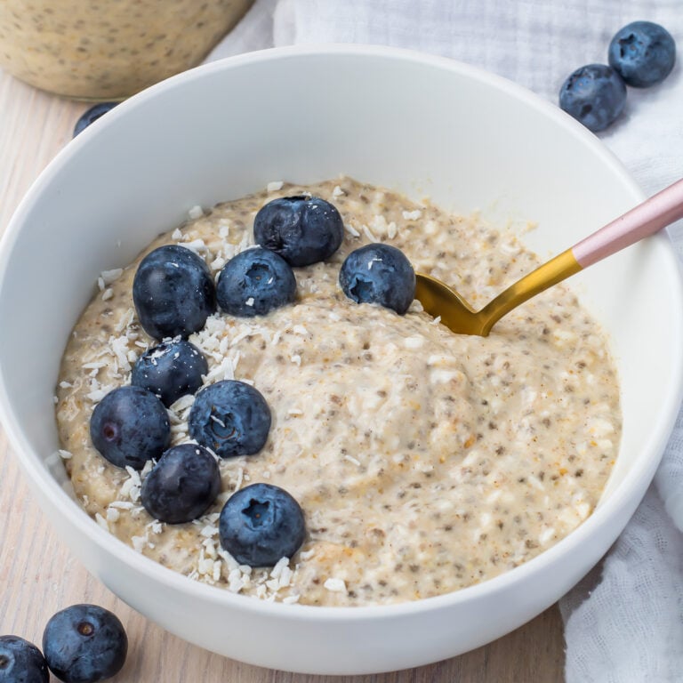 A bowl of keto overnight oats topped with fresh blueberries and shredded coconut, with a golden spoon resting inside. The bowl is placed on a light wooden surface with a white cloth nearby.
