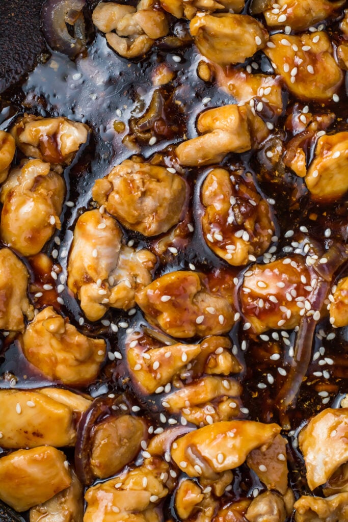 Close-up of cooked chicken pieces coated in a sticky teriyaki glaze, sprinkled with sesame seeds. The chicken is golden brown and shiny, highlighting the rich sauce.