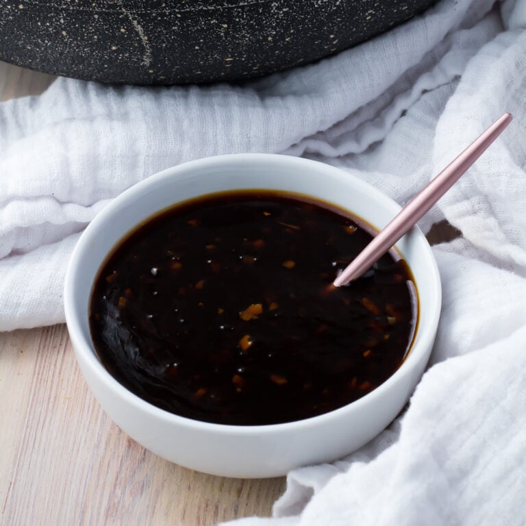 A white bowl filled with keto teriyaki sauce placed on a light wooden surface, accompanied by a metallic spoon. A white, textured cloth is draped around the bowl, adding contrast to the scene.