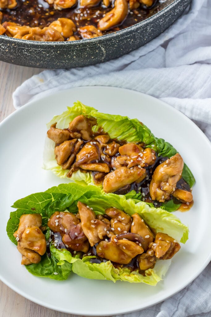 Teriyaki chicken lettuce wraps sprinkled with sesame seeds rest elegantly on a white plate, next to a pan on a crisp white tablecloth.