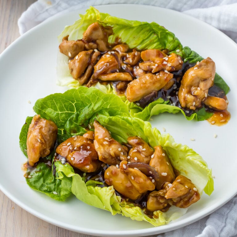 Two teriyaki chicken lettuce wraps filled with glazed chicken pieces, mushrooms, and garnished with sesame seeds are presented on a white plate. The dish is set on a light wooden surface with a white napkin in the background.