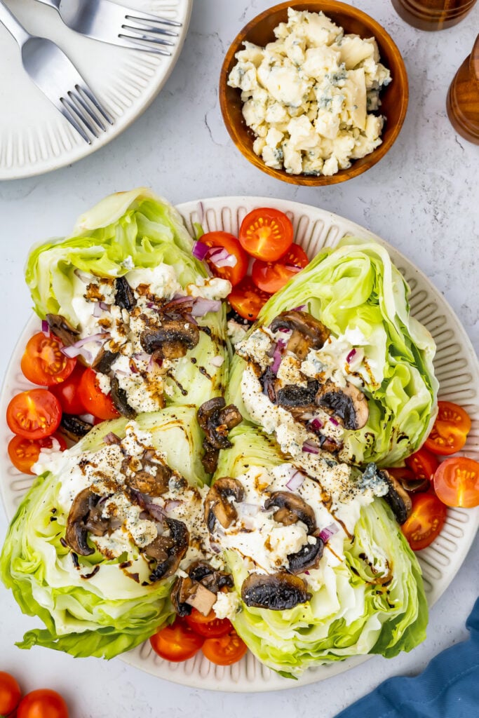 A wedge salad features four lettuce wedges topped with mushrooms, blue cheese, and red onions, arranged on a plate. Grape tomatoes surround the crisp wedges. A bowl with extra cheese sits beside cutlery and pepper shakers, completing this fresh presentation.