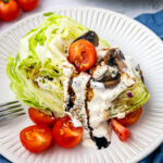 A wedge salad topped with creamy dressing, cherry tomatoes, sliced mushrooms, and crumbled cheese on a white plate. A fork rests beside the salad, with whole cherry tomatoes in the background.