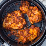 Cooked chicken pieces, seasoned with a reddish spice mix reminiscent of pollo asado, placed inside an air fryer basket. The chicken appears crispy and well-cooked.
