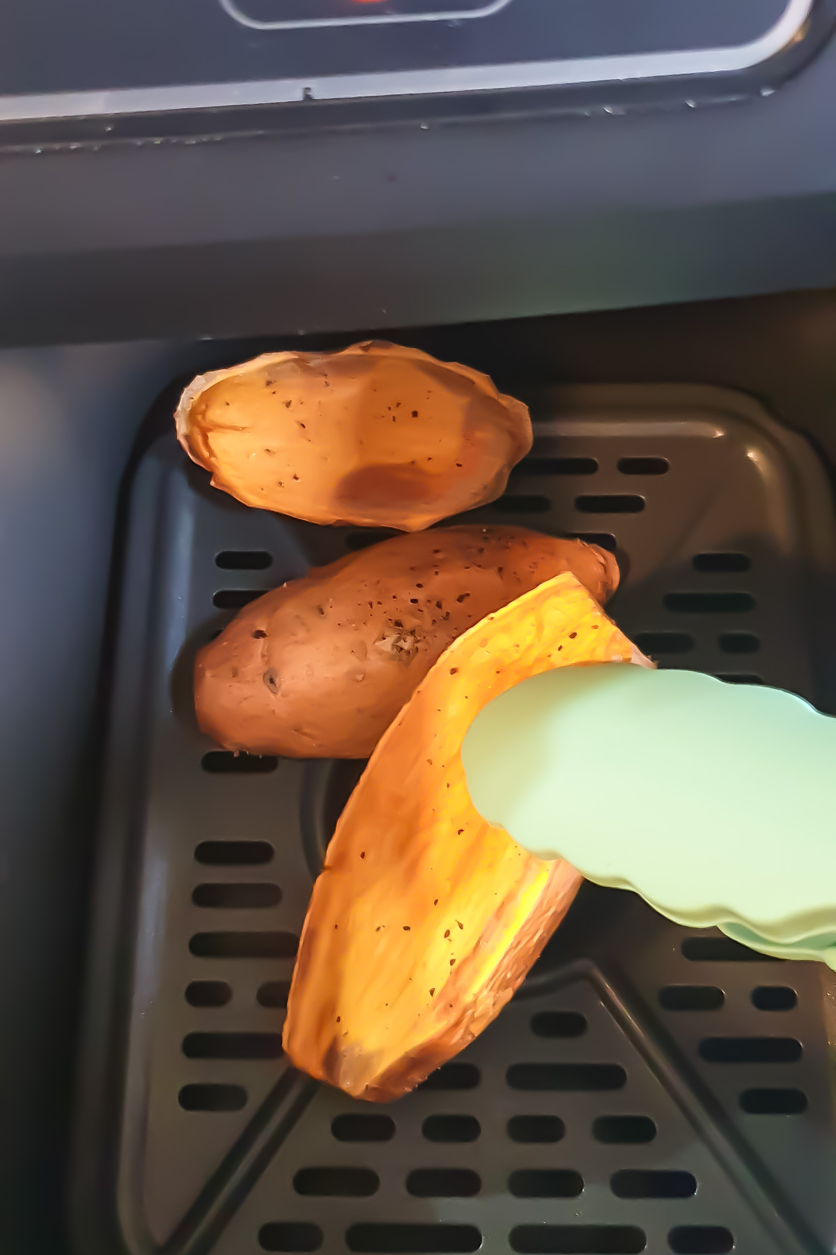A sweet potato cut in half, with one half being held by green tongs, sits inside an air fryer basket. The interior of the sweet potato is visibly cooked.