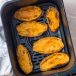 Five roasted sweet potato halves are arranged in an air fryer basket, their golden-brown, caramelized surfaces reminiscent of delectable sweet potato toast. The air fryer rests on a light wood surface next to a white cloth, ready to serve its tasty creation.