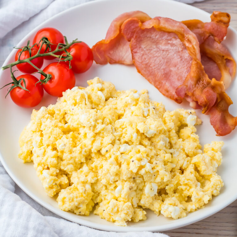 A plate with cottage cheese scrambled eggs, crispy bacon, and a cluster of fresh cherry tomatoes on a light wooden table.