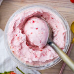 Close-up of a bowl of creamy pink keto strawberry-flavored ice cream with a scoop taken out. A spoon is placed beside the ninja creami pint. The table surface is light-colored, and there are small pieces of strawberries visible in the ice cream.