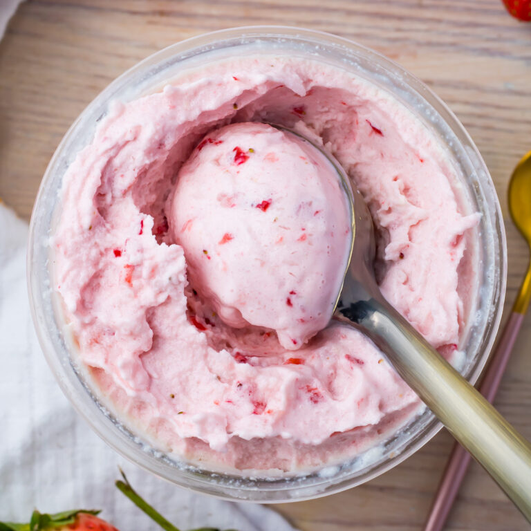 Close-up of a bowl of creamy pink keto strawberry-flavored ice cream with a scoop taken out. A spoon is placed beside the ninja creami pint. The table surface is light-colored, and there are small pieces of strawberries visible in the ice cream.