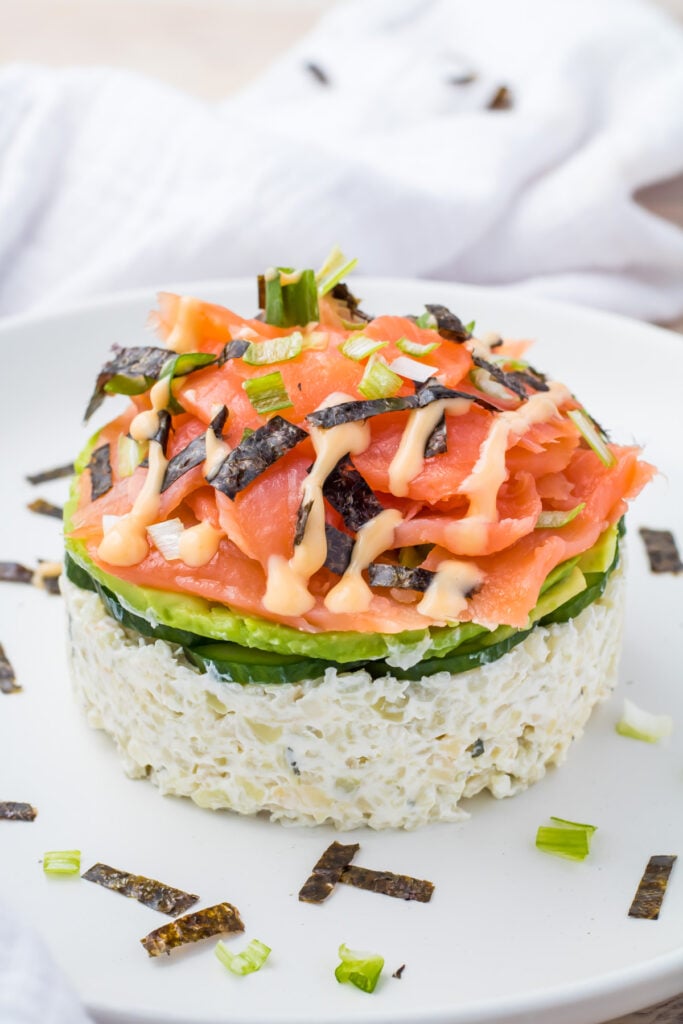 A delightful keto sushi stack with layers of sushi salad featuring a base of cauliflower rice, topped with crisp cucumber slices and savory smoked salmon, all drizzled in a creamy sauce. Sprinkled with nori pieces and chopped green onions, this dish is both visually stunning and deliciously satisfying.