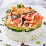 A sushi-inspired dish featuring a base of rice mixed with creamy sauce, topped with cucumber slices, avocado, raw salmon, and drizzled with sauce. Garnished with seaweed strips and chopped green onions.