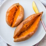 Two microwave baked sweet potatoes (halved) on a white plate with a gold knife. The potatoes are open-faced, revealing their vibrant orange interiors.