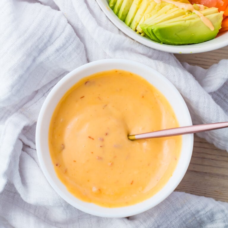 A bowl of creamy, spicy mayo without sriracha sits invitingly, with a spoon resting in it on a light, textured cloth. In the background, there's a partially visible bowl with sliced avocado.