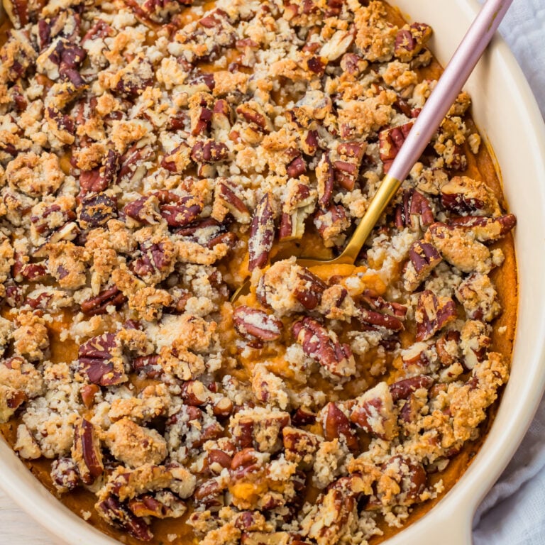 Close-up of a baked sweet potato apple casserole topped with a crumbly pecan streusel in a white oval dish. A spoon is partially submerged, revealing the golden-orange filling with hints of apple beneath the crispy topping.