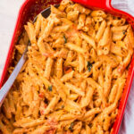 A red baking dish filled with creamy baked feta pasta mixed with cherry tomatoes and herbs. A serving spoon is placed inside the dish, ready to serve.