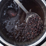 Cooked black beans in an instant pot with black liquid, being stirred by a slotted spoon.
