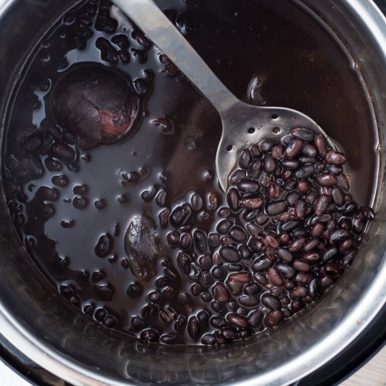 Cooked black beans in an instant pot with black liquid, being stirred by a slotted spoon.