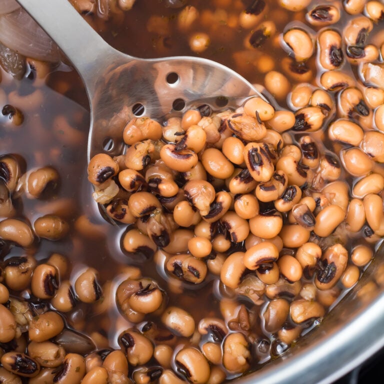 A stainless steel spoon lifting cooked black-eyed peas from an instant pot filled with more peas and cooking liquid. The beans are a beige color with distinctive black markings, and some liquid is visible.