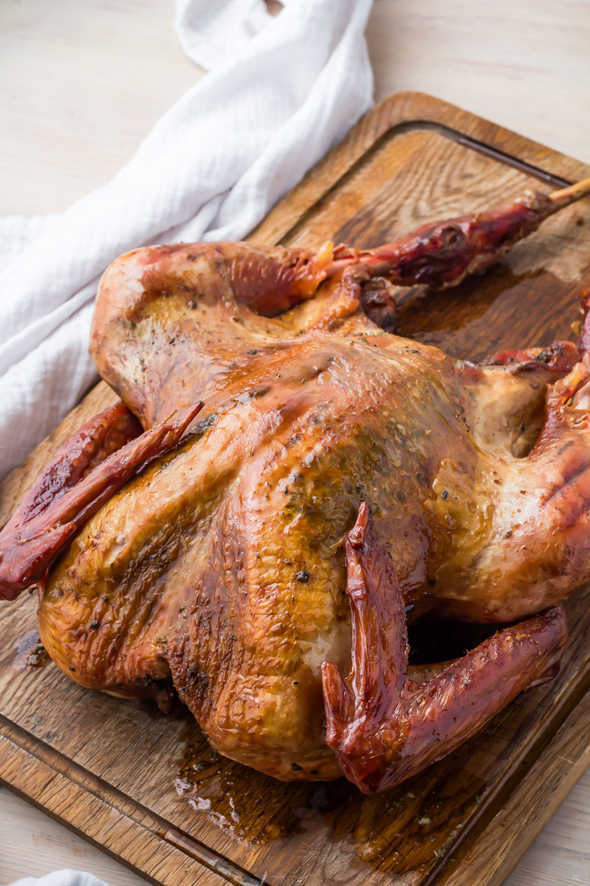 A roasted turkey, golden brown and crispy, rests on a wooden cutting board. A white cloth is partially visible in the background.