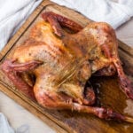Smoked turkey with golden brown skin resting on a wooden cutting board. The turkey is seasoned and has crispy skin, with white cloth napkins partially visible in the background.