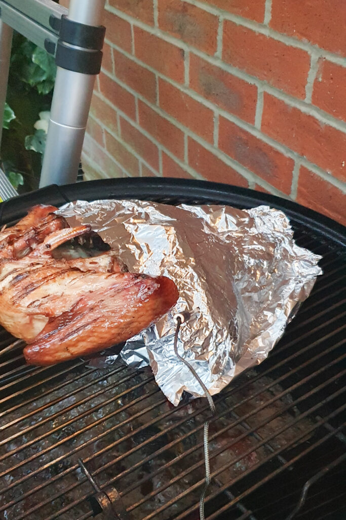 Smoked spatchcock turkey partially wrapped in aluminum foil, sits on a barbecue grill. The grill is positioned against a red brick wall. A metal probe is inserted into the meat. A garden ladder is visible in the background.
