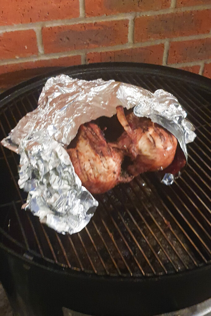 A smoked turkey on a grill is partially wrapped in crumpled aluminum foil. Set against a background of a brick wall.