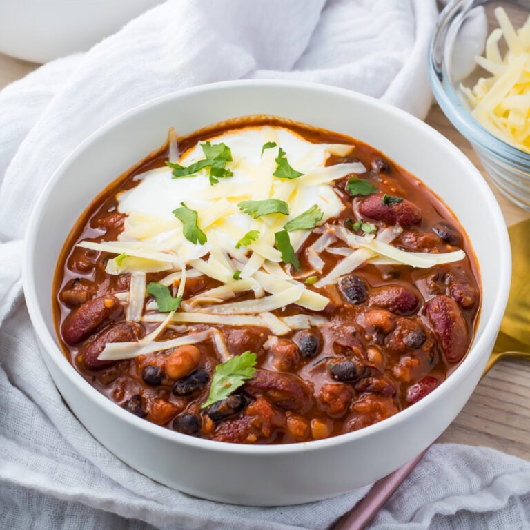 A bowl of vegan three bean chili featuring kidney, black, and pinto beans is topped with shredded cheese, sour cream, and cilantro. The bowl rests on a white cloth with a fork and a small dish of cheese nearby.