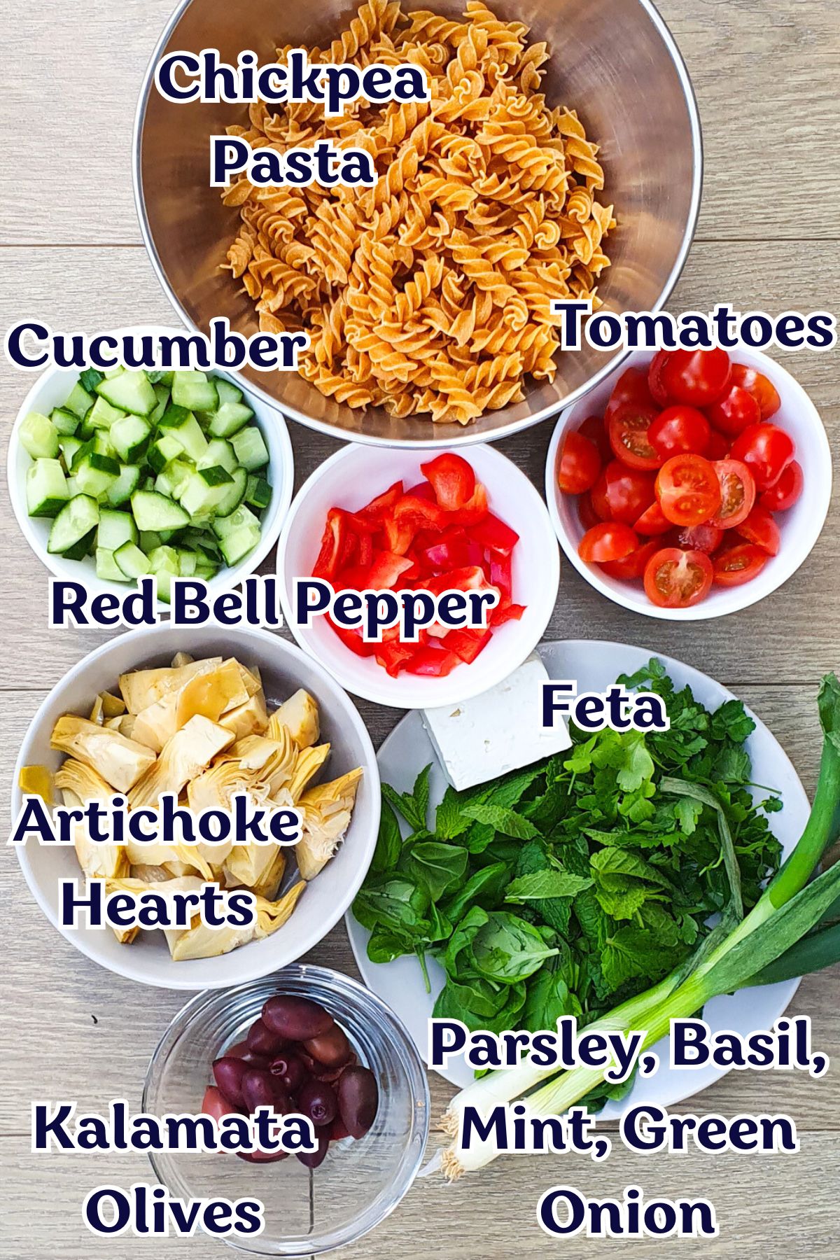 Bowls on a table contain ingredients for a salad: chickpea pasta, cherry tomatoes, cucumber, red bell pepper, feta cheese, artichoke hearts, parsley, basil, mint, green onion, and Kalamata olives. Each bowl is labeled.