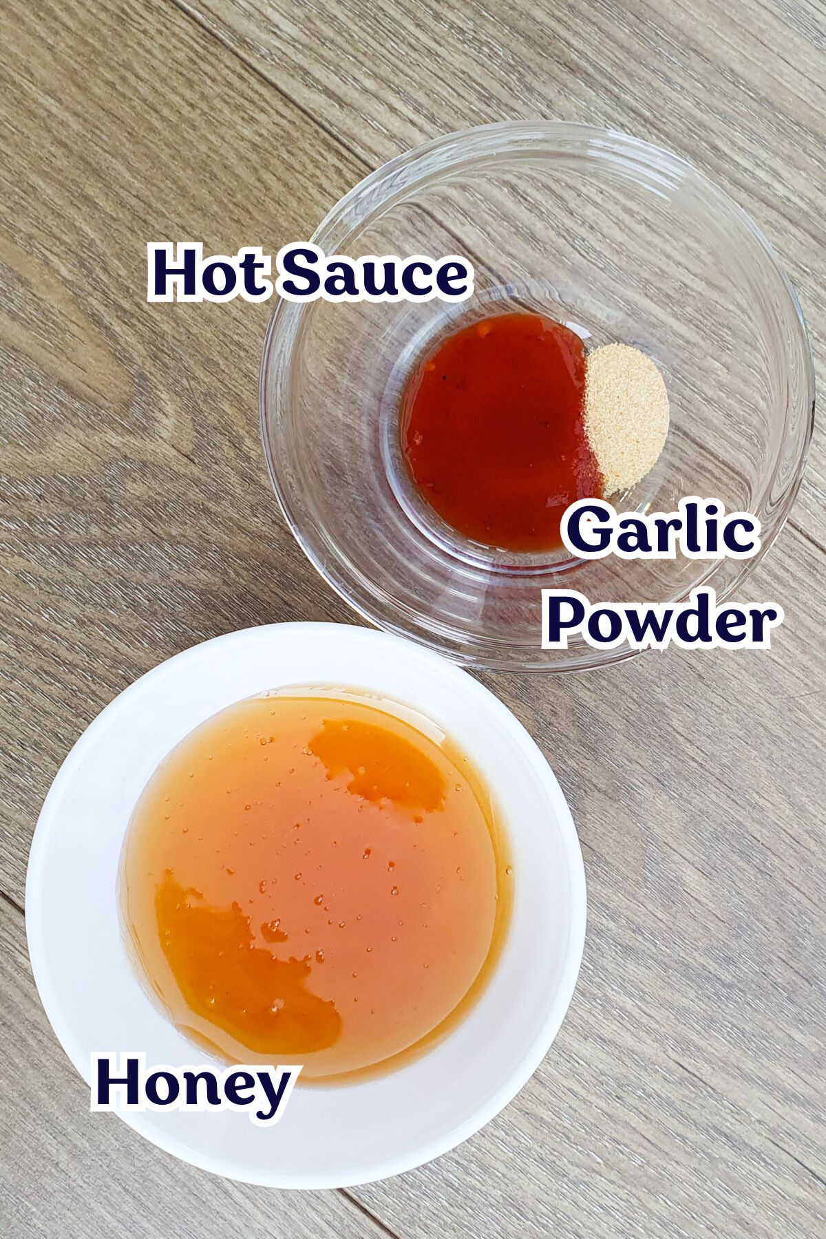 Two bowls with labeled ingredients on a wooden surface. The top bowl with hot sauce and garlic powder, and the bottom bowl contains honey.