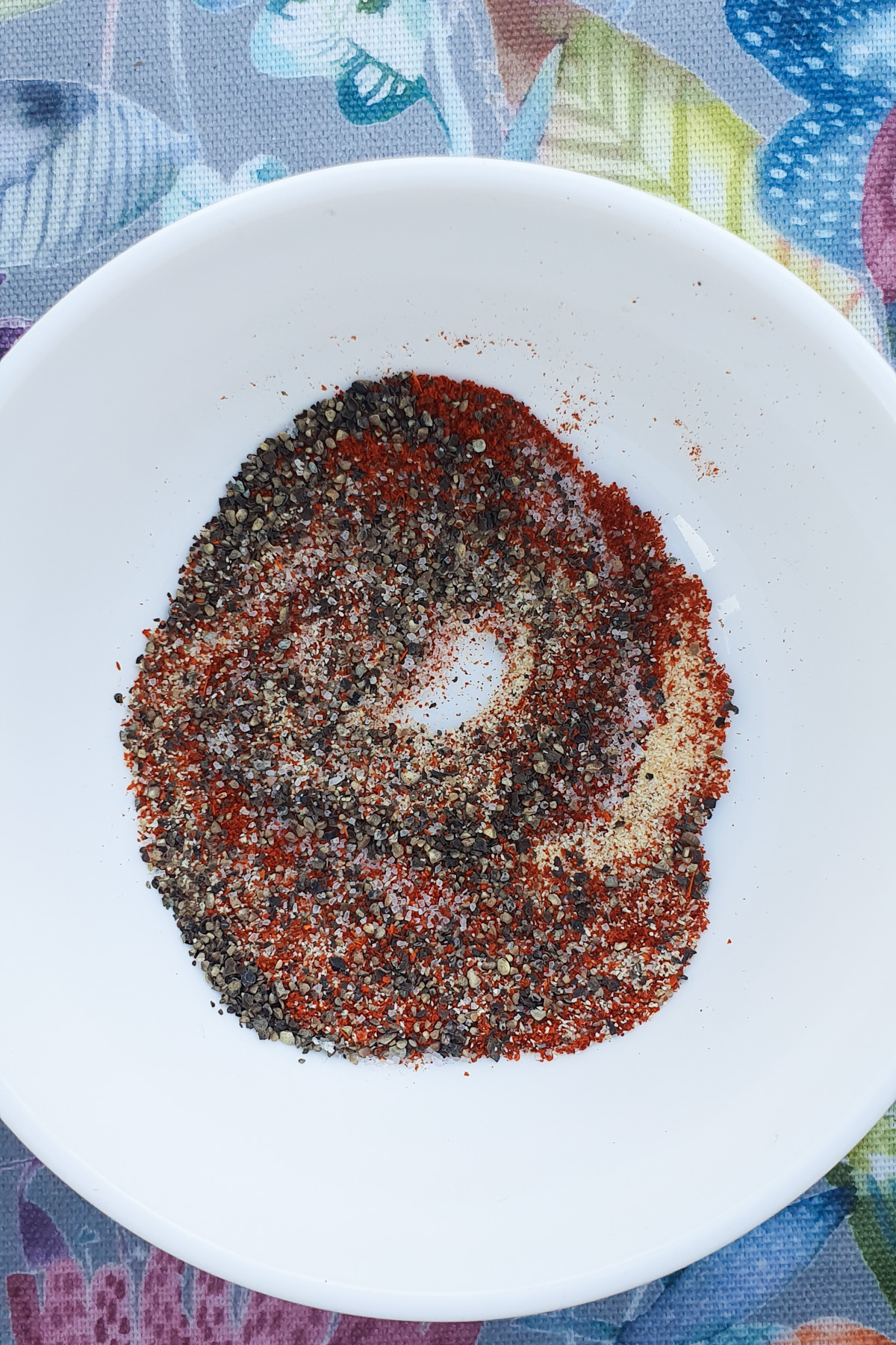 A white bowl containing a blend of ground spices arranged in a circular pattern. The bowl is placed on a colorful fabric background.