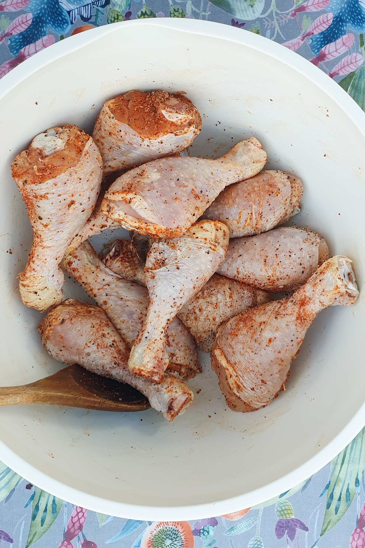A white bowl containing several raw chicken drumsticks seasoned with spices. A wooden spoon rests beside the drumsticks. The bowl is placed on a colorful floral-patterned tablecloth.