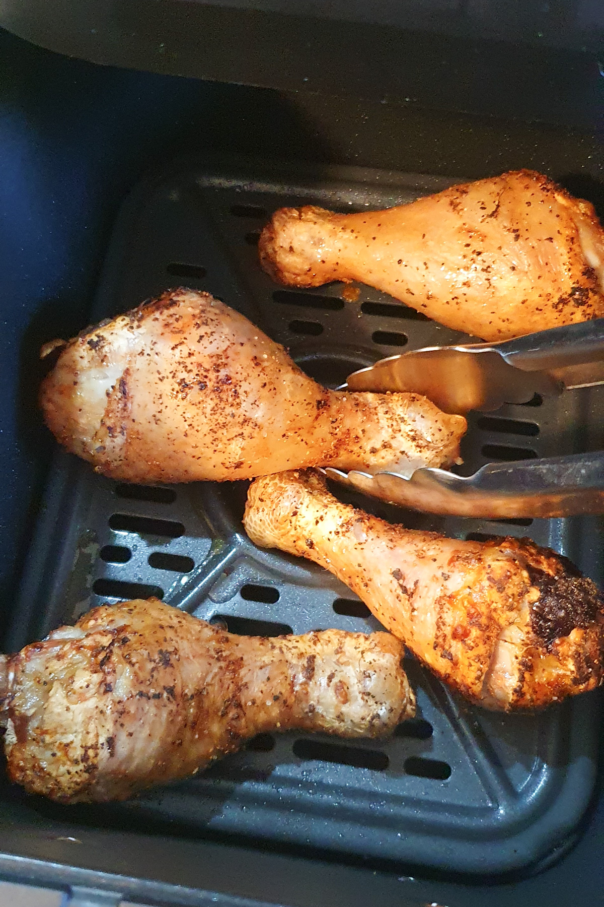 Four seasoned chicken drumsticks in an air fryer basket, with metal tongs holding one piece. The chicken appears cooked and slightly browned.