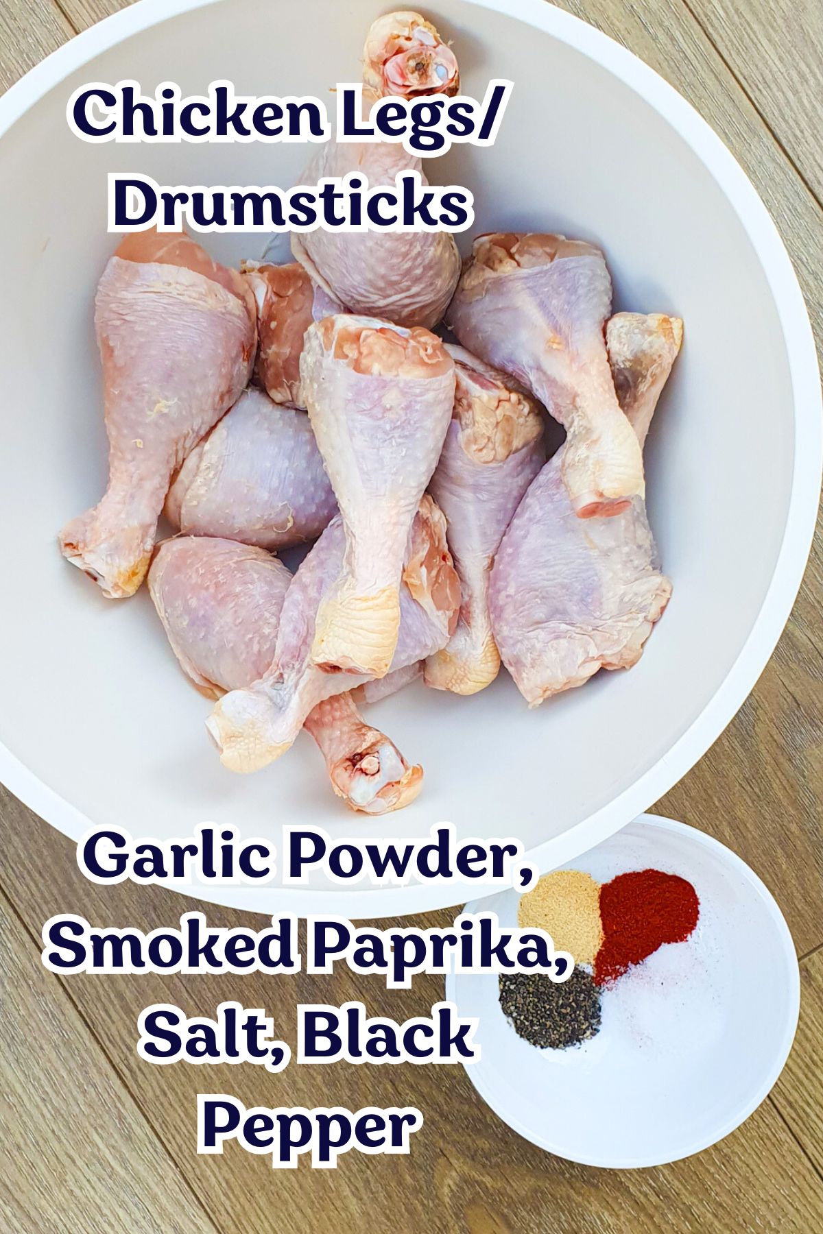 A bowl filled with raw chicken drumsticks. Next to it, a small bowl contains garlic powder, smoked paprika, salt, and black pepper. Text overlay lists "Chicken Legs/Drumsticks" and the seasonings.