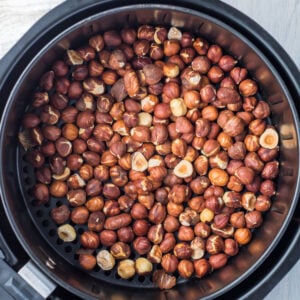 An overhead view of a large batch of air fryer hazelnuts being roasted inside an air fryer basket. The nuts are in various shades of brown, filling the circular basket to the brim.