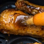 Close-up of a brush applying hot honey sauce to a seasoned and cooked chicken drumstick in an air fryer, showcasing the glistening texture and rich color of the meat.