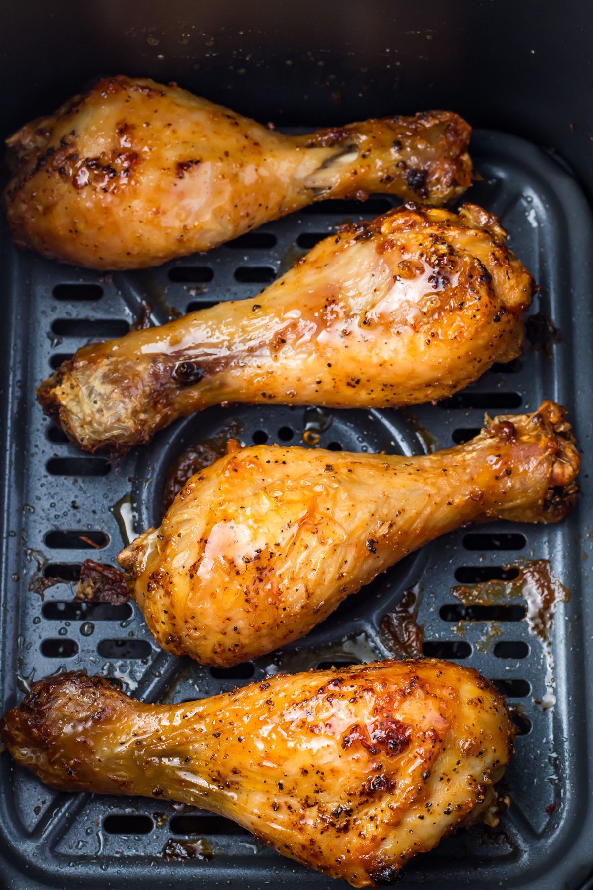 Four air fryer hot honey chicken drumsticks seasoned and cooked in an air fryer, lying on a dark perforated tray.