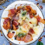 A baked camembert with bacon, basil, and a glaze of honey. The camembert is partially melted, surrounded by a sprinkle of red pepper flakes on a light blue surface.