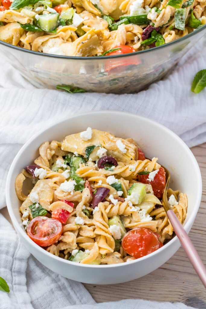 A bowl of pasta salad on a light-colored cloth. The salad includes fusilli pasta, cherry tomatoes, cucumbers, olives, greens, and crumbled cheese. A glass dish with more salad is in the background. A fork is resting in the bowl.