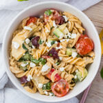 A bowl of gluten free pasta salad featuring rotini pasta, cherry tomatoes, cucumber pieces, artichoke hearts, black olives, and spinach, all topped with crumbled feta cheese. The dish is arranged on a wooden table with a white napkin nearby.