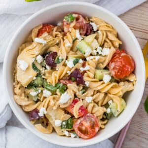 A bowl of gluten free pasta salad featuring rotini pasta, cherry tomatoes, cucumber pieces, artichoke hearts, black olives, and spinach, all topped with crumbled feta cheese. The dish is arranged on a wooden table with a white napkin nearby.